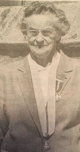 Catherine Trigwell, an older woman in glasses and a jacket, wearing a medal on a ribbon round her neck