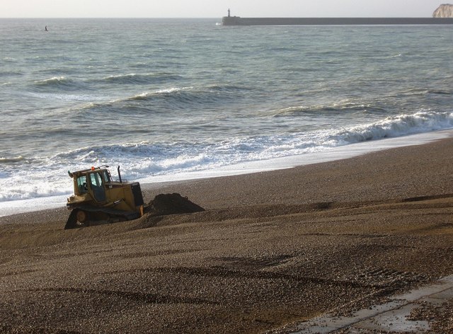 A bulldozer pushing shingle back up Seaford beach, with the Newhaven breakwater light on the horizon